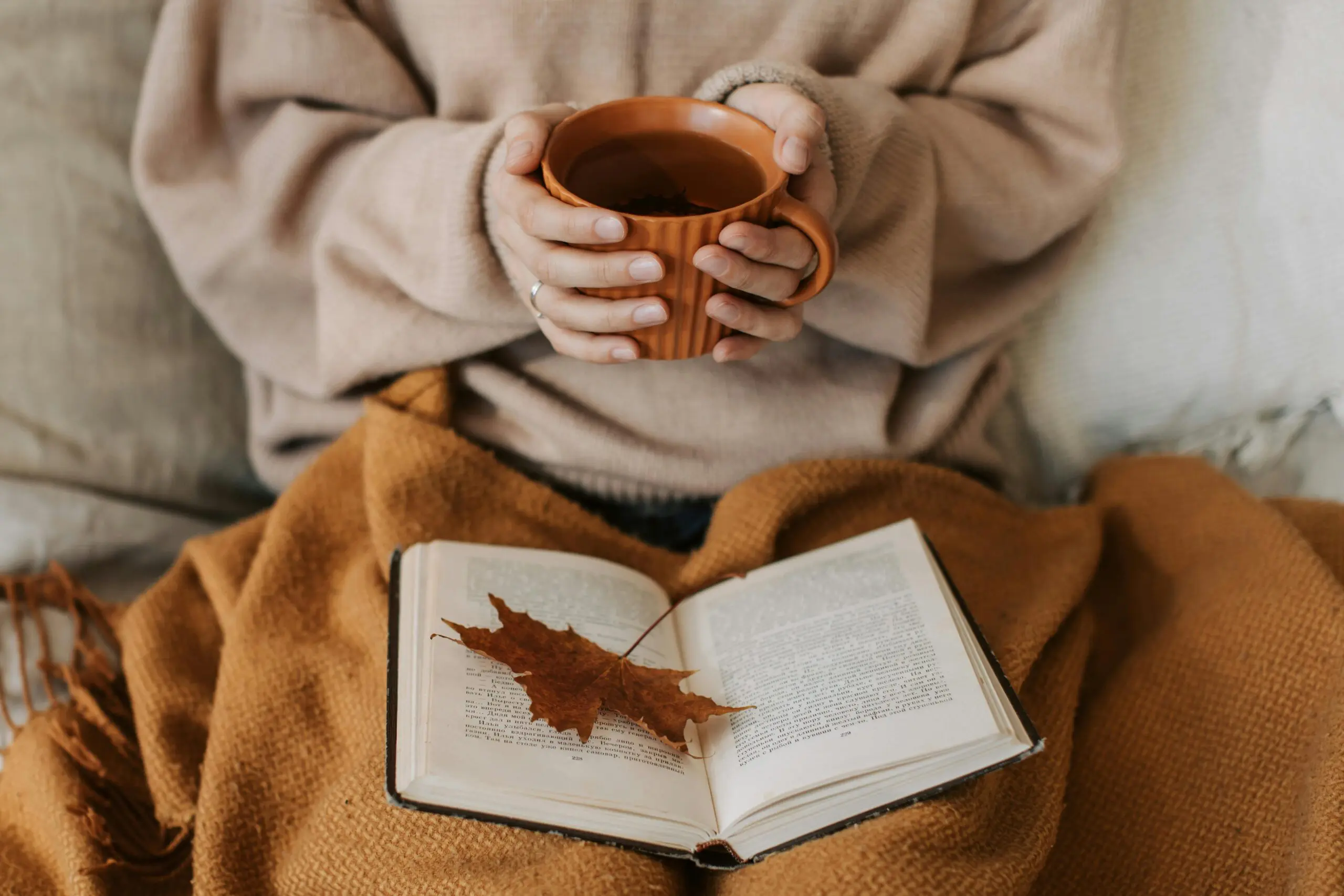 Woman reading a book with a coffee in hand under a brown blanket