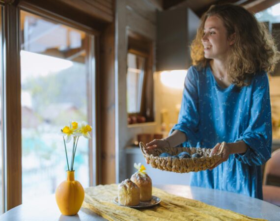 A smiling woman placing a basket on a dining table that has a plate of bread and a vase with flowers on it