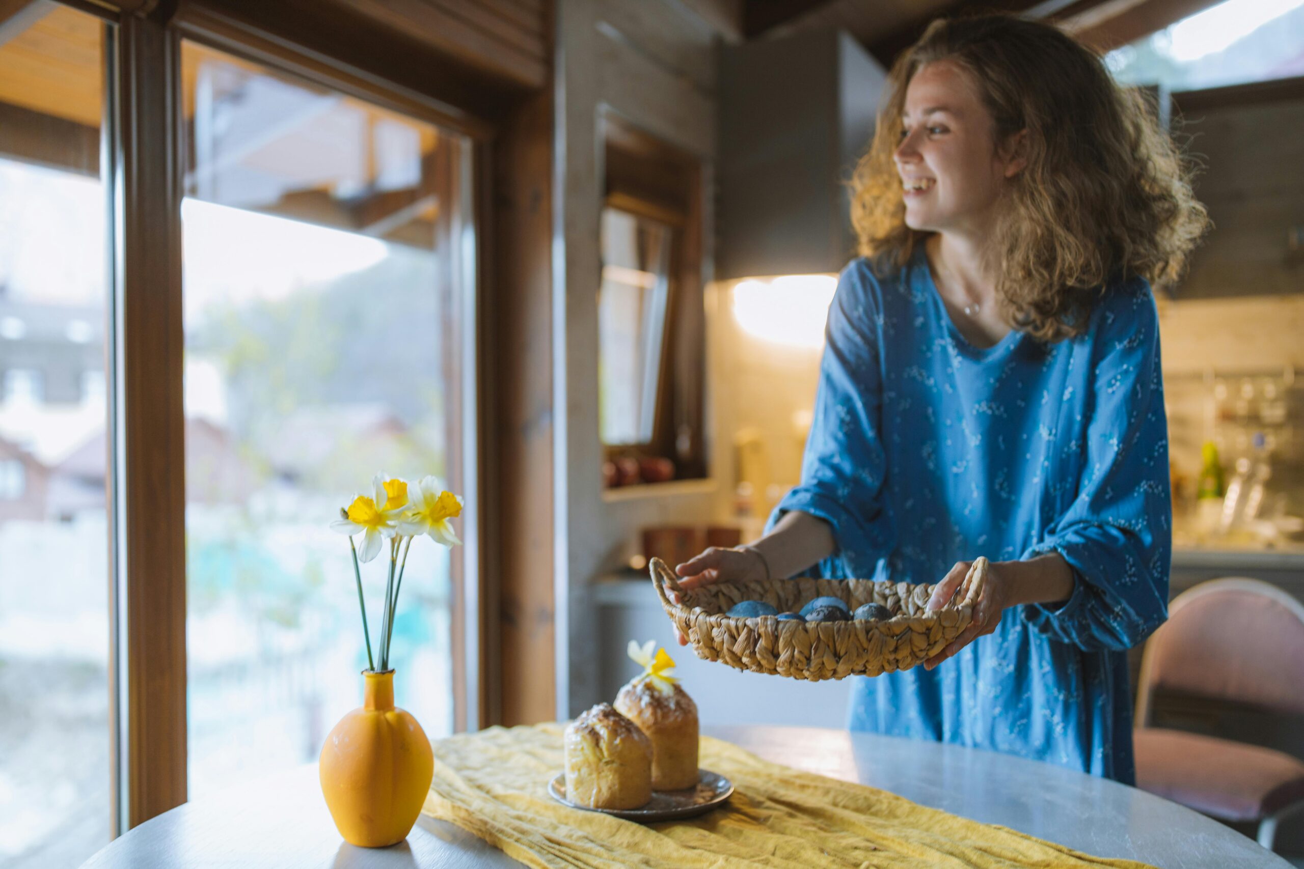 A smiling woman placing a basket on a dining table that has a plate of bread and a vase with flowers on it