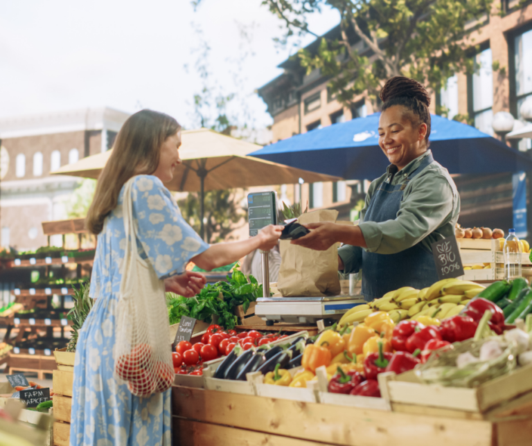 A woman buying vegetables from another woman at an outdoor farmers market booth